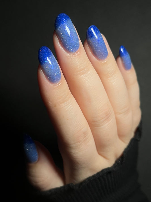 Hand with light blue to dark royal blue thermal nail polish on a dark background. Thermal nail polish with reflective glitter. Opal Theory Polish Boomerang Nebula.